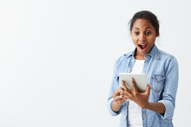 Porträt der aufgeregten jungen afroamerikanischen Frau, die vor Schock und Erstaunen schreit und neue Tablette in ihren Händen hält. Überraschtes, dunkelhäutiges Mädchen mit Käferaugen, das beeindruckt aussieht, kann ihren eigenen Augen nicht trauen