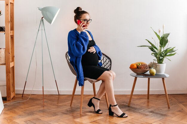 Porträt der bezaubernden Dame im schwarzen Kleid und in der blauen Strickjacke, die am Telefon sprechen und auf Holzstuhl sitzen. Schwangere Frau in Brille berührt Bauch.