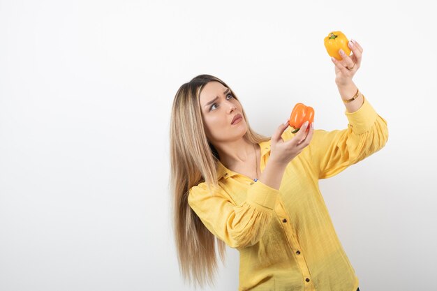 Porträt der blonden Frau, die bunte Paprika über weißer Wand betrachtet.