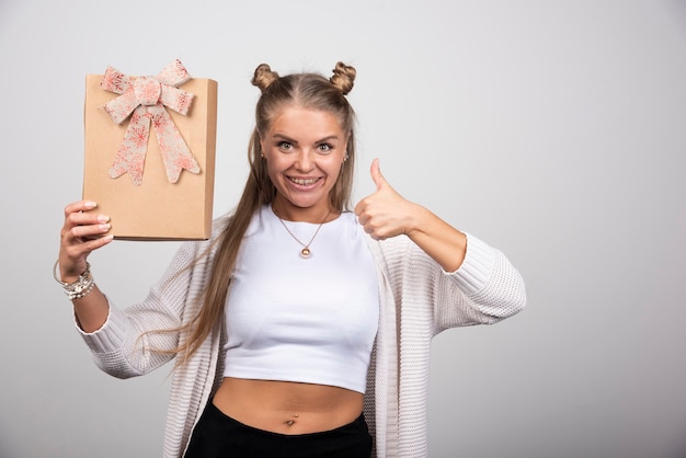 Porträt der blonden Frau, die Geschenkbox hält und Daumen aufgibt.