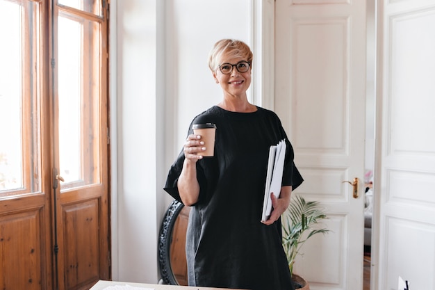 Porträt der blonden frau in brille und schwarzem kleid, das teetasse und dokumente hält