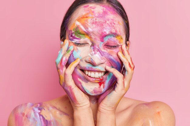 Porträt der brünetten jungen asiatischen Frau lächelt angenehm hält beide Hände auf Wangen steht mit geschlossenen Augen hat Gesicht von bunten Ölfarben verschmiert