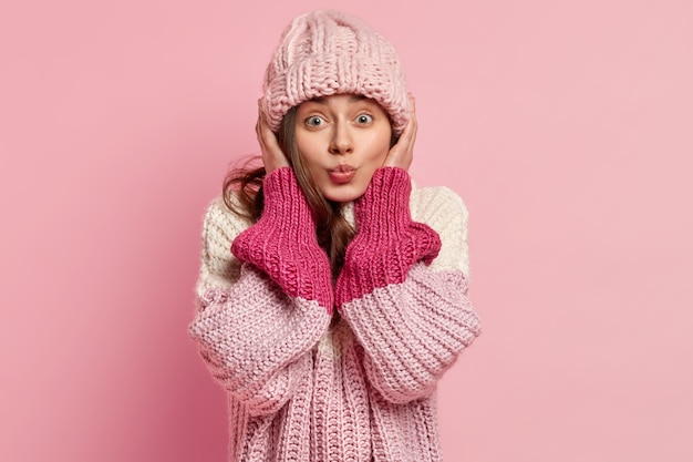 Porträt der europäischen Frau trägt gestrickte Kopfbedeckung, gekleidet in losen Pullover, will jemanden küssen, isoliert über rosa Wand. Lässiger Kleidungsstil. Wintermode-Konzept. Menschliche Mimik
