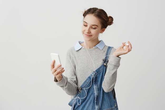 Porträt der Frau 20s, die auf Bildschirm des Handys mit angenehmem breitem Lächeln schaut. Charmanter weiblicher Teenager, der Selfie-Porträt macht, während Musik draußen hört. Interaktionskonzept