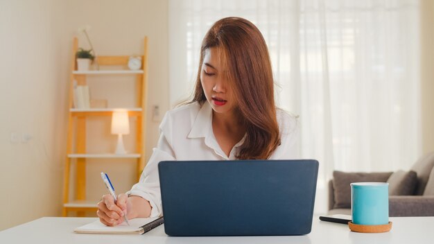 Porträt der freiberuflichen Freizeitkleidung der asiatischen Frauen unter Verwendung des Laptops, der im Wohnzimmer zu Hause arbeitet. Arbeiten von zu Hause aus, Fernarbeit, Selbstisolierung, soziale Distanzierung, Quarantäne zur Vorbeugung von Coronaviren.