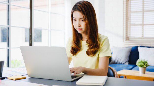 Porträt der freiberuflichen Freizeitkleidung der asiatischen Frauen unter Verwendung des Laptops, der im Wohnzimmer zu Hause arbeitet. Arbeiten von zu Hause aus, Fernarbeit, Selbstisolierung, soziale Distanzierung, Quarantäne zur Vorbeugung von Koronaviren.