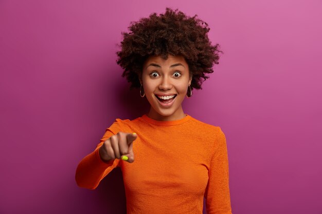 Porträt der freudigen dunkelhäutigen jungen Frau zeigt gerade mit dem Zeigefinger, sieht etwas Erstaunliches, sagt hey du, trägt orangefarbenen Pullover, isoliert über lebhafter lila Wand.