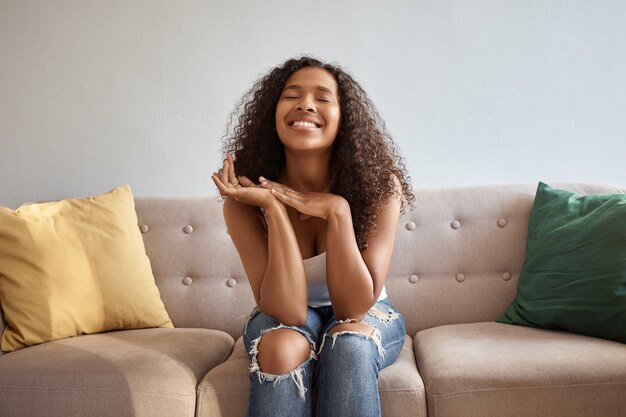 Porträt der freudigen emotionalen jungen dunkelhäutigen Frau, die im Wohnzimmer auf bequemer Couch aufwirft, in stilvollen zerlumpten Jeans sitzt, mit Händen unter ihrem Kinn aufblickt, in guter Stimmung ist, lächelt