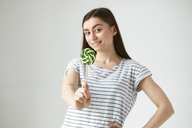 Porträt der freudigen schönen jungen Frau der Brünette im gestreiften lässigen T-Shirt, das Lutscher hält und mit positivem fröhlichem Gesichtsausdruck lächelt. Selektiver Fokus auf Bonbons