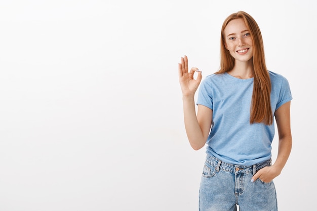 Porträt der freundlichen und freudigen gutaussehenden rothaarigen Frau mit Sommersprossen, die Hand in der Tasche halten, lässiges Gespräch, das sicherstellt, dass die Arbeit rechtzeitig erledigt wird und okay oder ausgezeichnete Geste zeigt