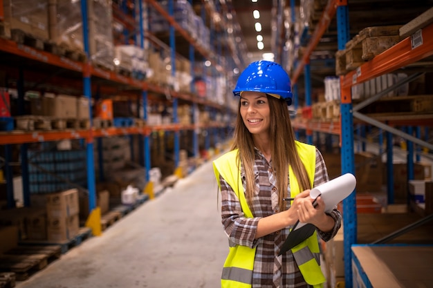 Porträt der fröhlichen Frau in der Schutzuniform, die Pakete und Lagerbestand der Produkte im Lagerraum überprüft