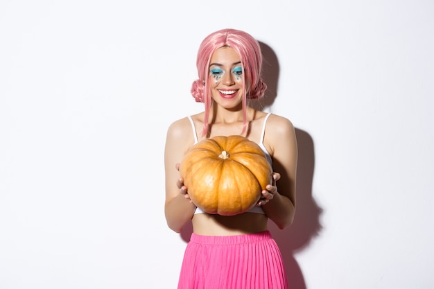 Porträt der fröhlichen Frau mit rosa Perücke und hellem Make-up, das Kürbis für Halloween glücklich betrachtet, stehend.
