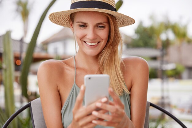 Porträt der glücklich entzückten Frau trägt Strohhut, hat fröhlichen Ausdruck, liest etwas auf dem Smartphone, genießt Freizeit in teurem Straßencafé, hat Sommerruhe. Lächelnde Frau mit Handy