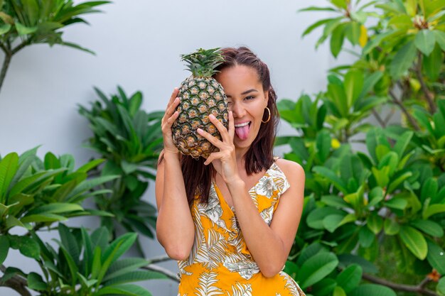 Porträt der glücklichen asiatischen lächelnden Frau im gelben Sommerkleid halten Ananas