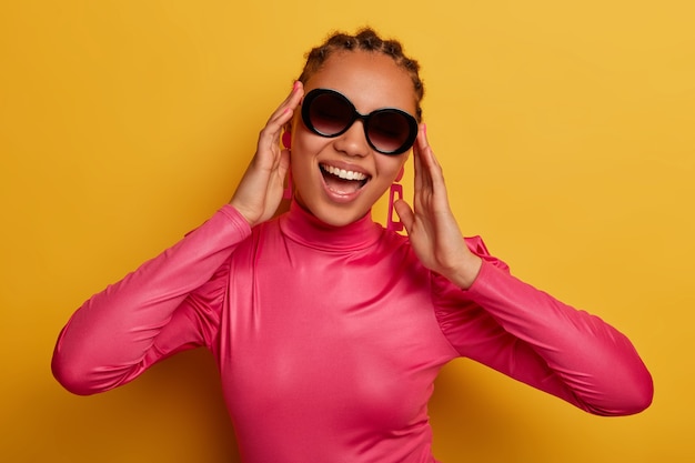 Porträt der glücklichen dunkelhäutigen jungen Frau trägt trendige Sonnenbrille und rosa Pullover, genießt sonnigen Tag, gekleidet in hellen Kleidern, isoliert auf gelber Wand. Menschen-, Mode- und Stilkonzept.