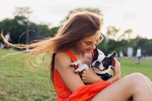 Porträt der glücklichen hübschen Frau, die auf Gras im Sommerpark sitzt, Boston-Terrier-Hund hält, positive Stimmung lächelt, orange Kleid trägt, trendigen Stil, Sonnenbrille, spielt mit Haustier