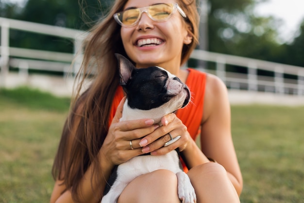 Kostenloses Foto porträt der glücklichen hübschen frau, die auf gras im sommerpark sitzt, boston-terrier-hund hält, positive stimmung lächelt, orange kleid trägt, trendigen stil, sonnenbrille, spielt mit haustier