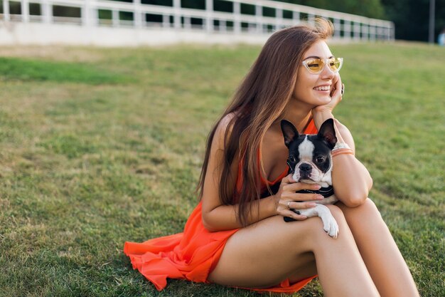 Porträt der glücklichen hübschen Frau, die auf Gras im Sommerpark sitzt, Boston-Terrier-Hund hält, positive Stimmung lächelt, orange Kleid trägt, trendigen Stil, Sonnenbrille, spielt mit Haustier
