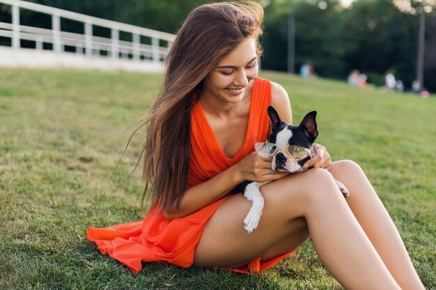 Porträt der glücklichen hübschen Frau, die auf Gras im Sommerpark sitzt, Boston-Terrier-Hund hält, positive Stimmung lächelt, orange Kleid trägt, trendigen Stil, Sonnenbrille, spielt mit Haustier