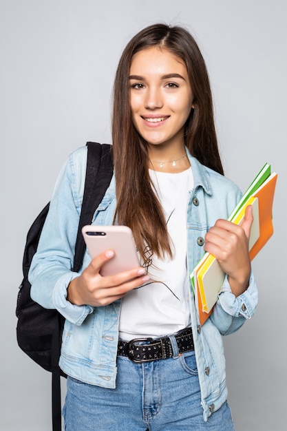 Porträt der glücklichen jungen Frau, die mit Rucksack hält Bücher und Handy lokalisiert auf weißer Wand steht