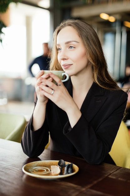 Porträt der glücklichen jungen Frau mit Becher in den Händen, die Kaffee am Morgen am Restaurant trinken