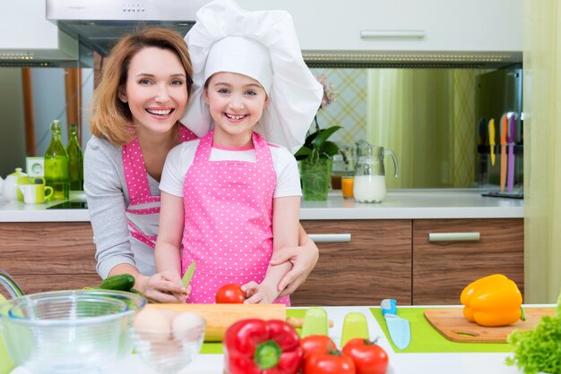 Porträt der glücklichen jungen Mutter mit Tochter in der rosa Schürze, die an der Küche kocht