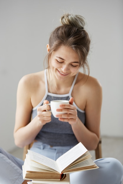 Porträt der glücklichen jungen schönen Frau, die lesendes Lesebuch hodierende Tasse Kaffee sitzt, der auf Boden über weißer Wand sitzt.