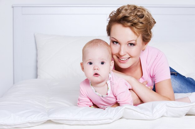 Porträt der glücklichen jungen schönen Mutter, die mit ihrem Baby auf dem Bett liegt