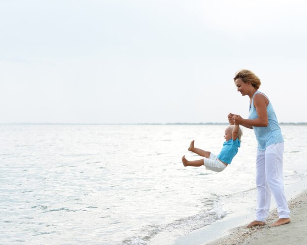 Porträt der glücklichen lächelnden jungen Mutter mit dem kleinen Kind, das am Strand spielt.