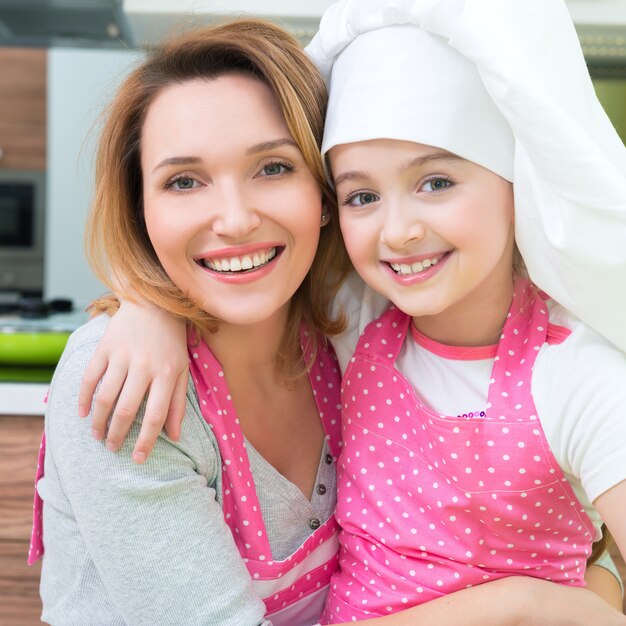 Porträt der glücklichen lächelnden Mutter und der Tochter in der rosa Schürze an der Küche