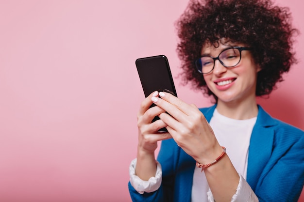 Porträt der glücklichen modernen Frau mit der kurzen Frisur gekleideten blauen Jacke, die Smartphone auf Rosa mit glücklichem Lächeln rollt