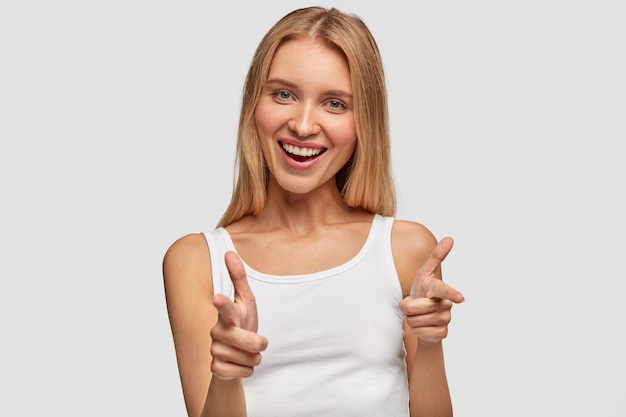 Porträt der glücklichen niedlichen blonden Frau mit freudigem Ausdruck, zeigt mit Vorderfingern auf Sie