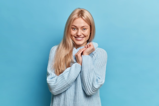 Porträt der glücklichen schüchternen jungen Frau hält Hände zusammen sieht positiv aus, trägt lässig gestrickten Pullover posiert gegen blaue Wand