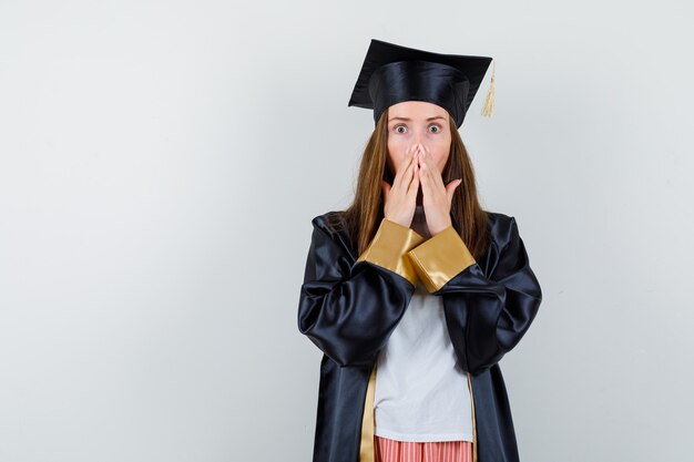 Porträt der graduierten Frau, die Hände auf Mund in lässiger Kleidung, Uniform hält und schockierte Vorderansicht schaut