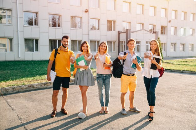 Porträt der Gruppe glücklicher Studenten in der zufälligen Ausstattung mit den Büchern, die sich Daumen zeigen