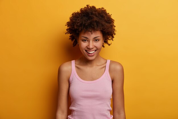 Porträt der gut aussehenden aufrichtigen Frau sieht positiv aus, hat zahniges Lächeln, direkten Blick, gesunde Haut, gekleidet in lässigem Outfit, isoliert über gelber Wand. Fröhliche Gefühle