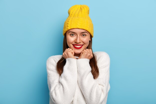 Porträt der gut aussehenden Frau sieht positiv aus, lächelt vor Vergnügen, trägt roten Lippenstift, hat dunkles Haar, gelben Hut, weißen Pullover