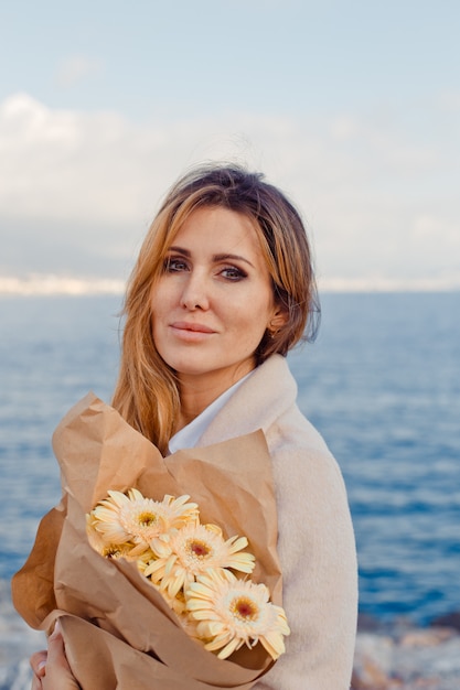 Porträt der hübschen Dame mit Blumen, die am Nachmittag am Meer stehen.