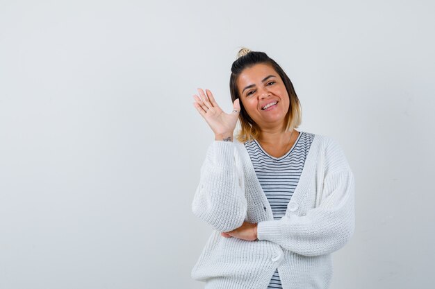 Porträt der hübschen Frau, die Hand zum Gruß im T-Shirt, in der Strickjacke und in der fröhlichen Vorderansicht winkt