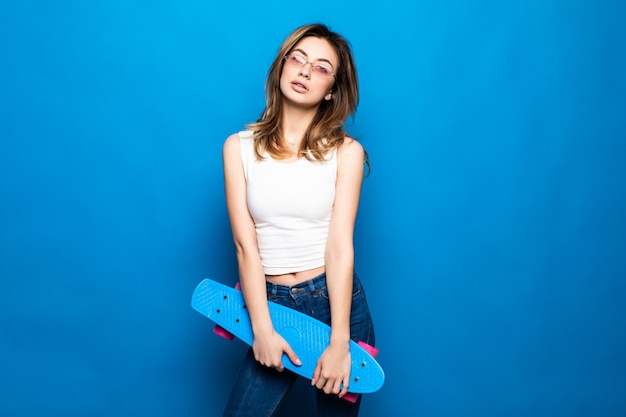 Porträt der hübschen jungen Frau in der Freizeitkleidung stehend, Skateboard lokalisiert auf blauer Wand haltend. Menschen Lifestyle-Konzept.