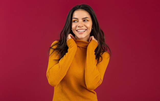 Porträt der hübschen lächelnden stilvollen brünetten hispanischen Frau im gelben Herbstwintermodekleidpullover, der lokalisiert auf roter Wand aufwirft