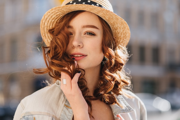 Porträt der interessierten Ingwerfrau, die im eleganten Hut aufwirft. Außenaufnahme des herrlichen weiblichen Modells mit rotem lockigem Haar, das Wochenende in der Stadt verbringt.