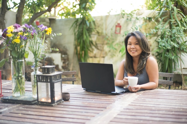 Porträt der japanischen Frau mit Laptop im Hinterhof. Schönes Mädchen mit braunen Haaren, das online einkaufen oder chatten, Spaß haben, Filme gucken, Freiberufler arbeiten. Kaffee trinken