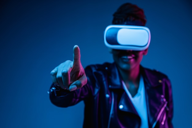 Porträt der jungen Afroamerikanerfrau, die in VR-Brille im Neonlicht auf blauem Hintergrund spielt
