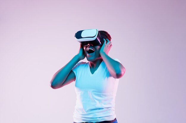 Porträt der jungen Afroamerikanerfrau, die in VR-Brille im Neonlicht auf Gradientenhintergrund spielt. Konzept menschlicher Emotionen, Gesichtsausdruck, moderner Geräte und Technologien. Sieh erstaunt aus.