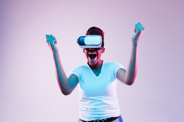 Porträt der jungen Afroamerikanerfrau, die in VR-Brille im Neonlicht auf Gradientenhintergrund spielt