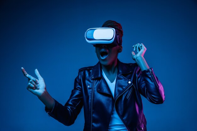 Porträt der jungen afroamerikanischen Frau, die in VR-Brille im Neonlicht auf Blau spielt.