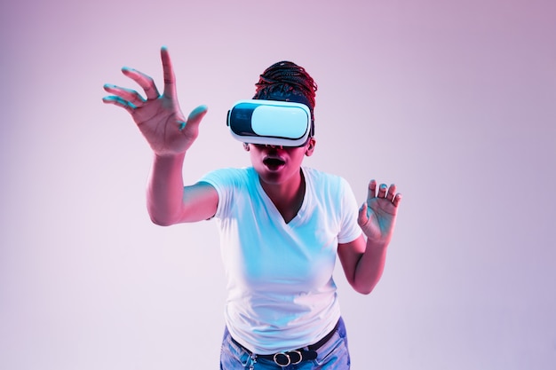 Porträt der jungen afroamerikanischen Frau, die in VR-Brille im Neonlicht auf Gradient spielt.
