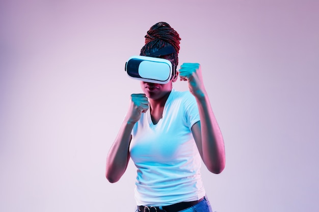 Porträt der jungen afroamerikanischen Frau, die in VR-Brille im Neonlicht auf Gradient spielt.