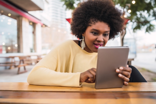 Porträt der jungen afroamerikanischen lateinamerikanischen Frau, die ihr digitales Tablett beim Sitzen am Coffeeshop verwendet. Technologiekonzept.